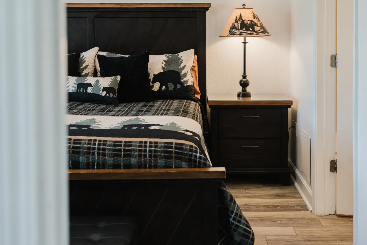 This inviting bedroom features a bed adorned with cozy bear-themed bedding, a soft glow from a table lamp with a bear motif, and a wooden nightstand. The décor is warm and welcoming, providing a sense of comfort and rustic charm.