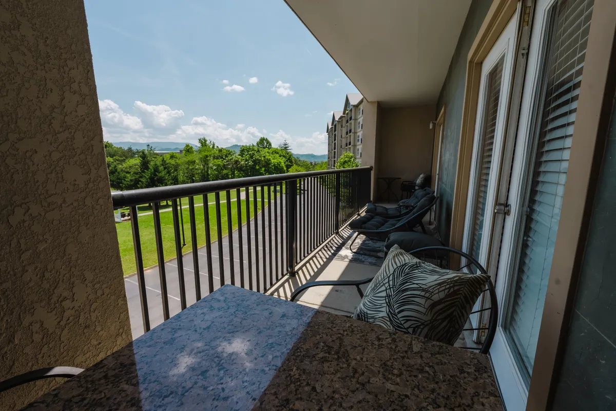 A serene private patio area featuring a balcony railing, two comfortable chairs, and a granite-topped table, all overlooking lush greenery. The scene captures the tranquility of early morning or dusk, ideal for relaxing with a book, enjoying coffee, or having conversations while taking in beautiful mountain views.