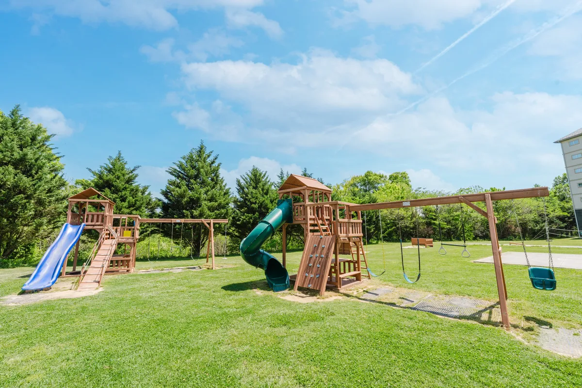 A vibrant playground with swings and a slide sits on a well-kept green lawn, inviting children to play while families gather for picnics or relax outdoors. The scene is framed by lush grass and a friendly, community-oriented atmosphere.