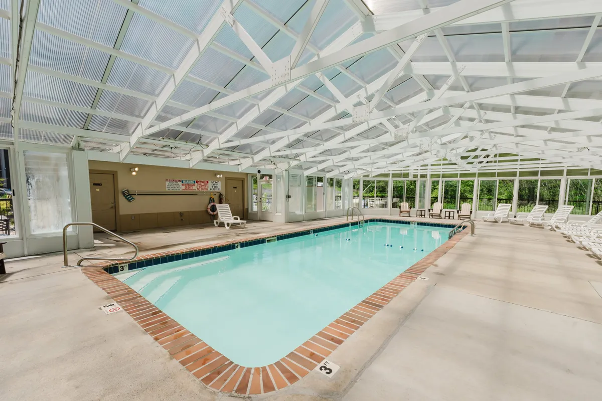 A bright indoor swimming pool area with a glass roof and comfortable lounge chairs lining the pool. The space feels both refreshing and relaxing, ideal for unwinding or enjoying time with loved ones regardless of the weather outside.