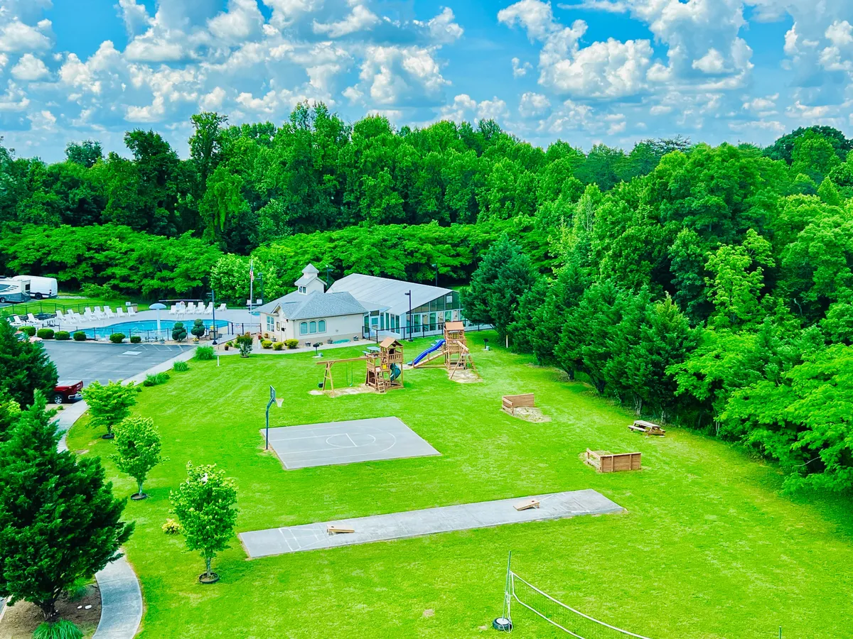 An exterior view that combines playground equipment, a basketball court, a swimming pool, lush green trees, and a community building. The scene is lively, showing various activities that appeal to all ages.