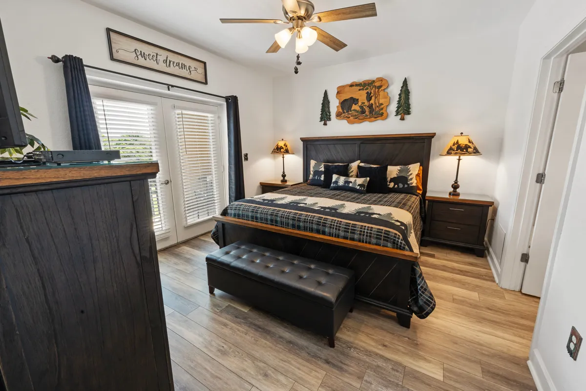 A king-sized bed sits at the heart of a tranquil bedroom, flanked by wooden nightstands with table lamps. Above the bed hangs decorative wall art, and curtains frame the window, allowing gentle natural light. The space exudes calm and comfort, inviting restful sleep or quiet moments with a book.