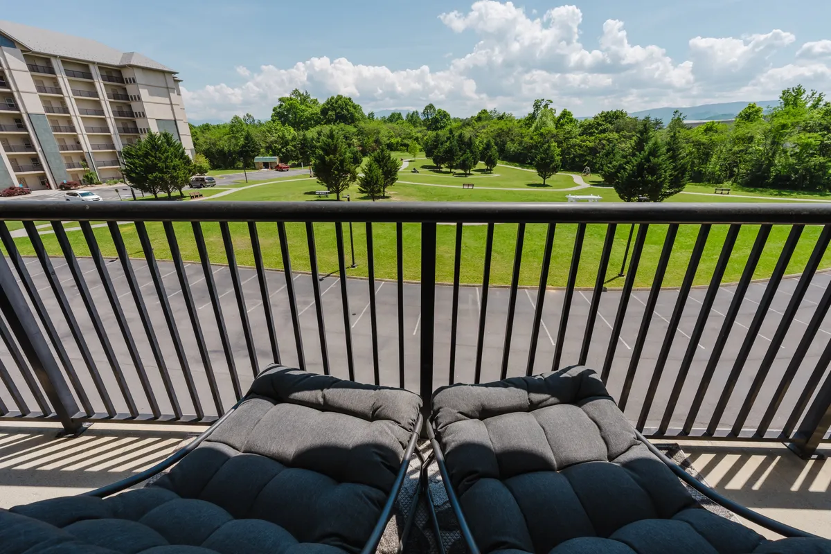 A tranquil balcony scene featuring comfy chairs overlooking a green lawn and leafy trees, with a cloudy sky overhead. The set-up suggests a perfect spot for relaxing, reading, or gazing at mountain views from the comfort of your own space.