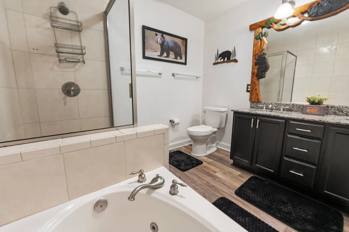 A cozy bathroom features a spacious jacuzzi tub set under calming bear-themed artwork, with granite countertops and dark cabinetry completing the soothing environment. This inviting space is designed for relaxation, offering a private spot to unwind after a busy day.